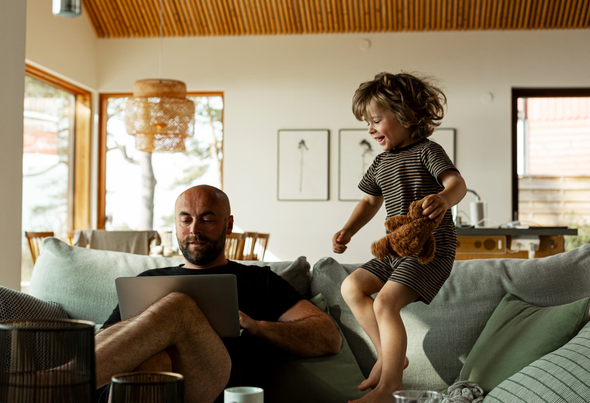 Pappa med dator i soffan medan ett barn hoppar med en nalle i ett ljust vardagsrum